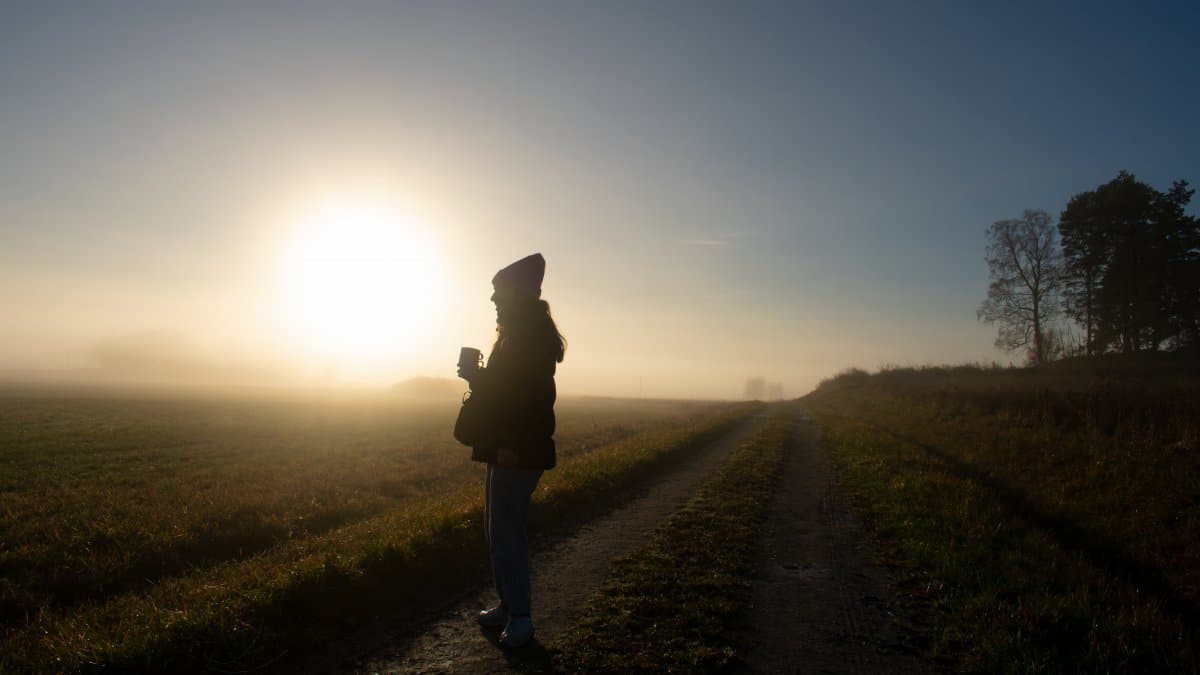 Silhouette of person holding a cup on a rural path with a sunrise over the misty fields in Sweden.