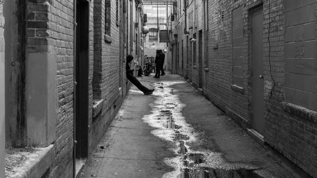 Atmospheric black and white photograph of a narrow alley with puddles and people.