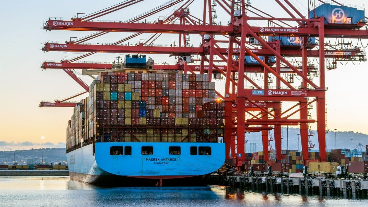 A large container ship is docked at a bustling port with red cranes loading cargo.