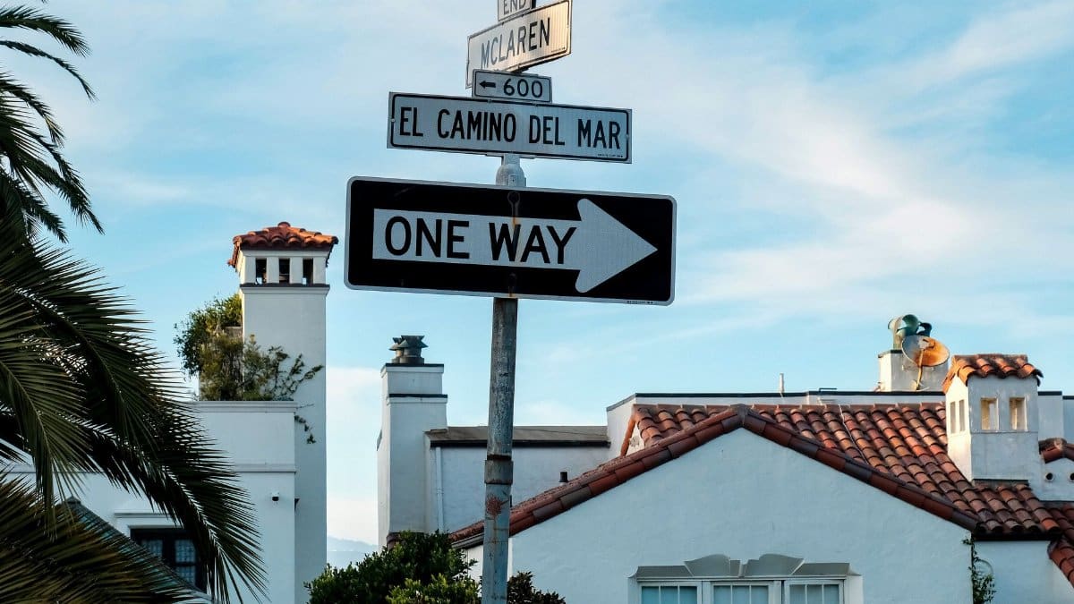 Street signs with Spanish-style architecture, depicting direction and local street names on a sunny day.