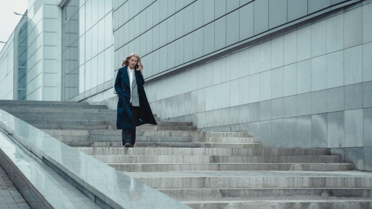 Confident woman in formal coat walking downstairs and talking on her smartphone outdoors.