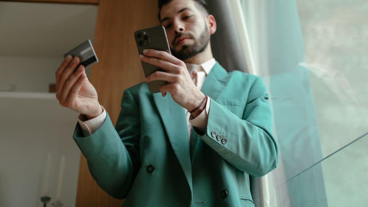 Man in teal blazer using smartphone and credit card for online shopping indoors.