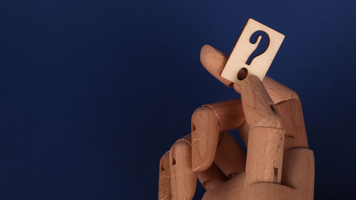 Wooden mannequin hand holding a block with a question mark against a blue background.