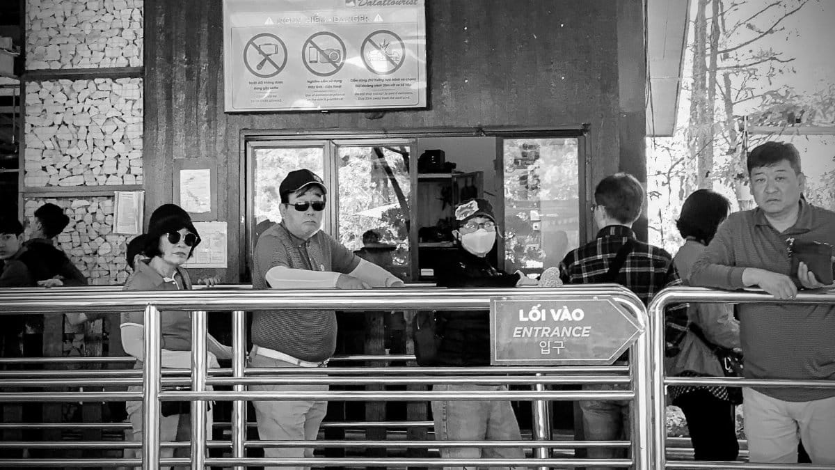 People waiting in line at an entrance with safety signage, captured in monochrome.