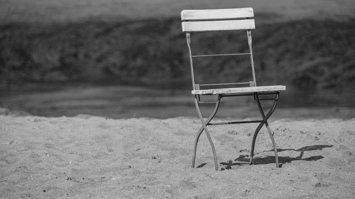 An empty wooden chair sits on a sandy beach, captured in black and white, evoking solitude.