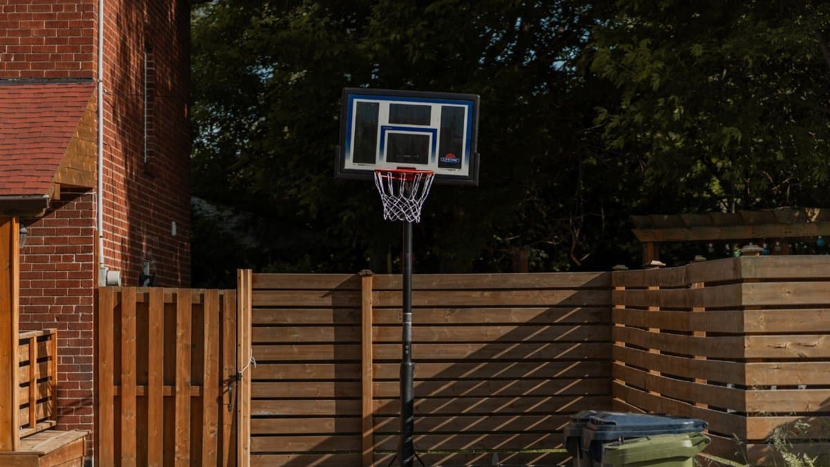 A backyard basketball hoop stands beside a brick house and wooden fence, ideal for outdoor sports.