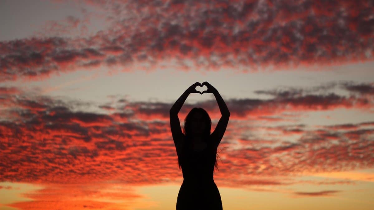 A silhouette of a woman creating a heart shape with her hands against a vibrant sunset.