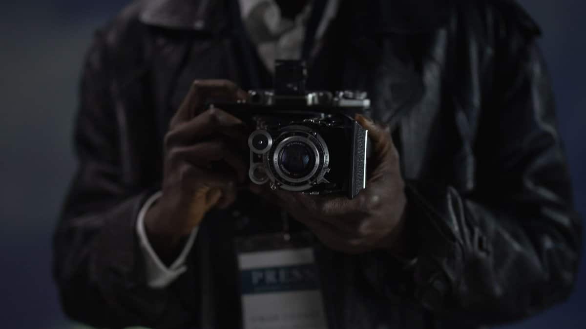 A close-up photograph of a person holding a vintage camera, possibly a reporter.