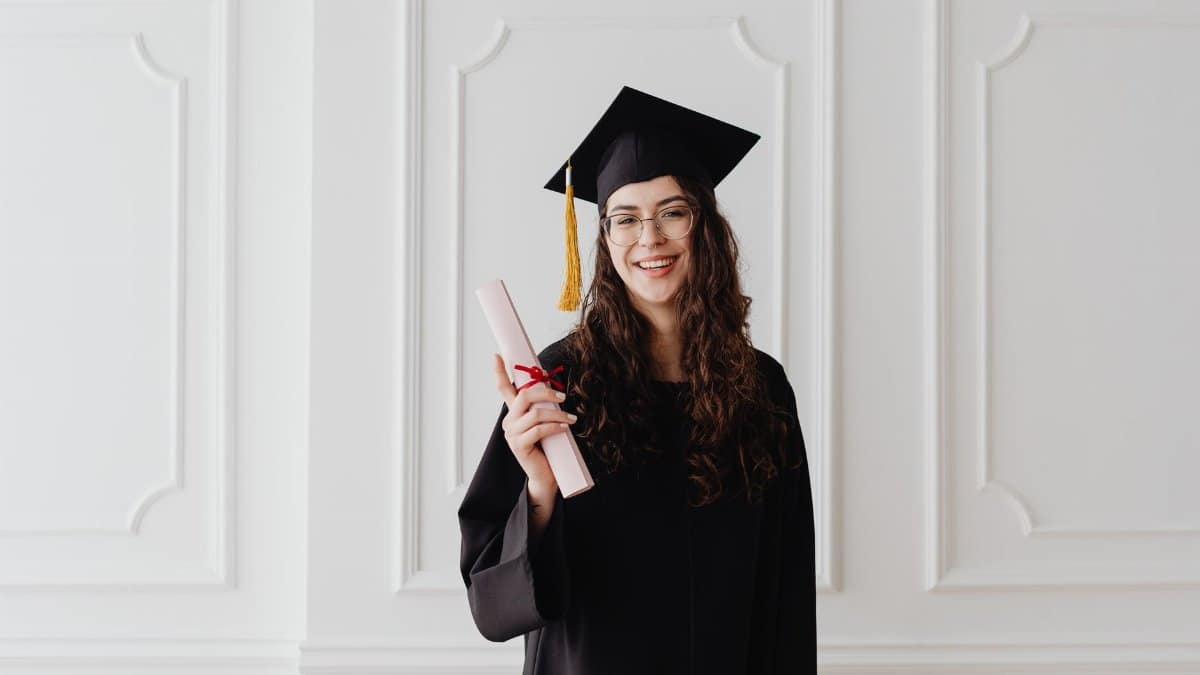 Smiling young woman in graduation gown and cap holding a diploma indoors.