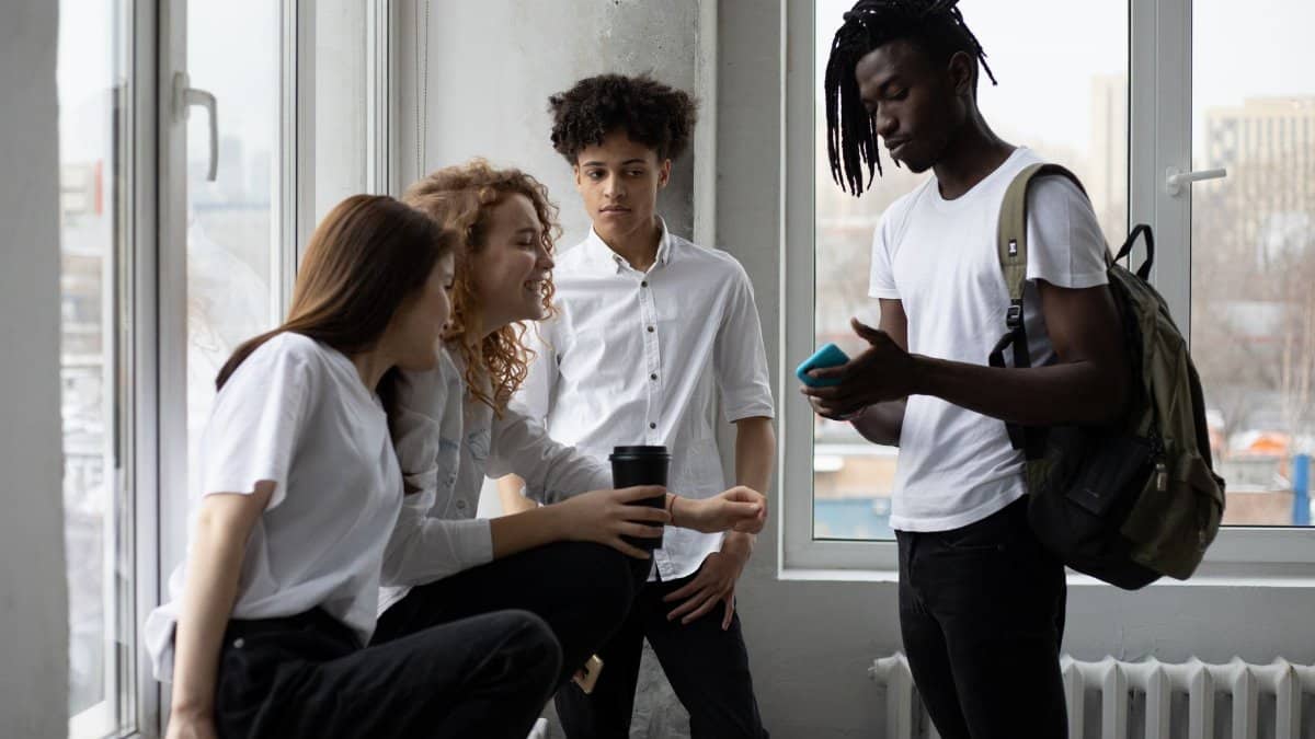 Group of multiracial classmates with backpack and takeaway coffee having conversation at windowsill near window in light classroom before lesson
