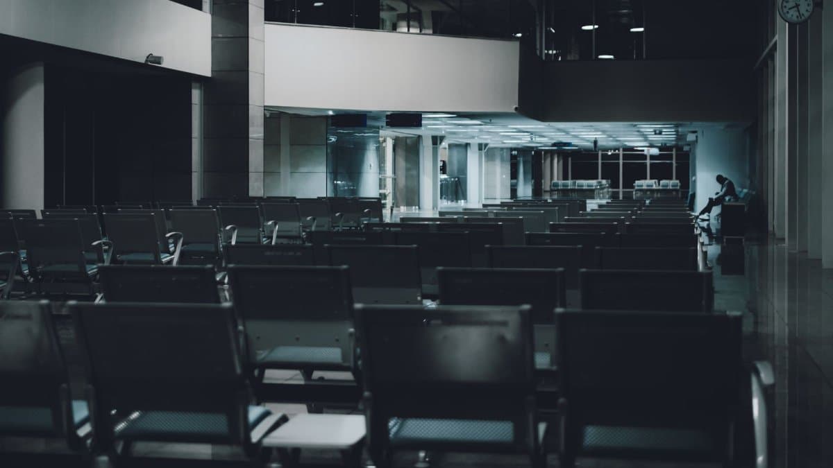 A quiet, dimly lit airport waiting area with unoccupied rows and a lone figure in the distance.