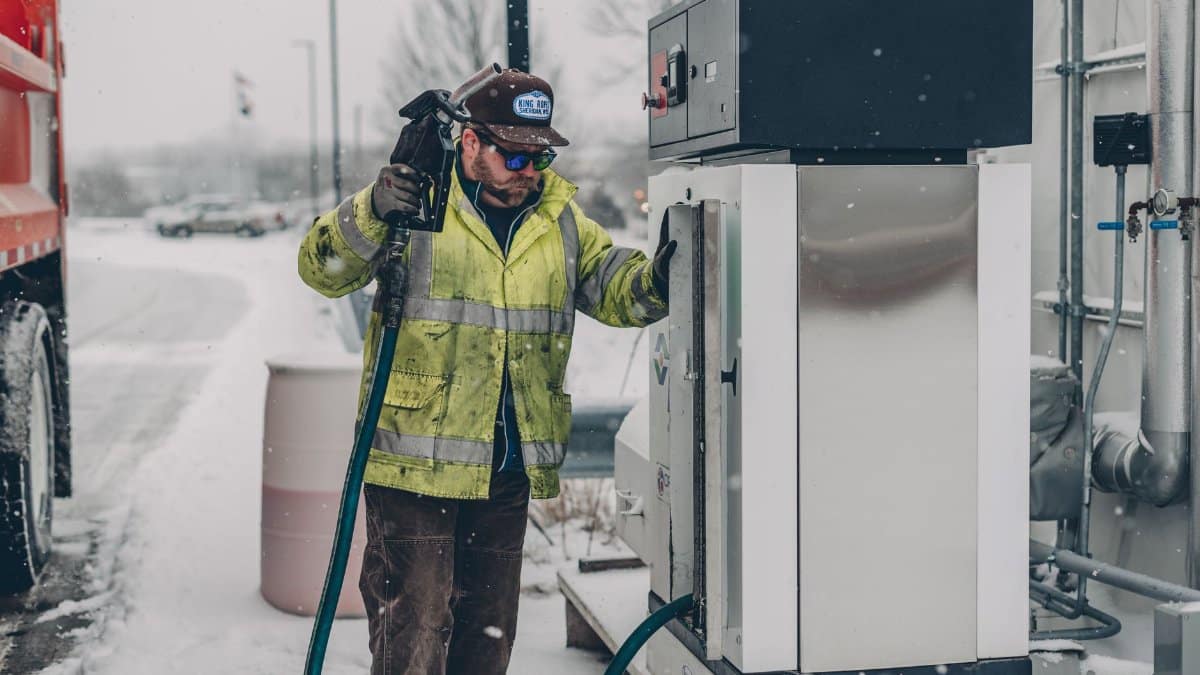 A man wearing a jacket refuels a truck at a gas station during winter with snow falling.
