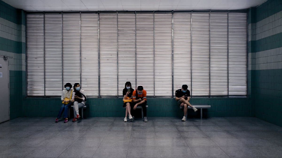 Full body of unrecognizable people in medical masks sitting on benches in spacious hall of clinic against windows
