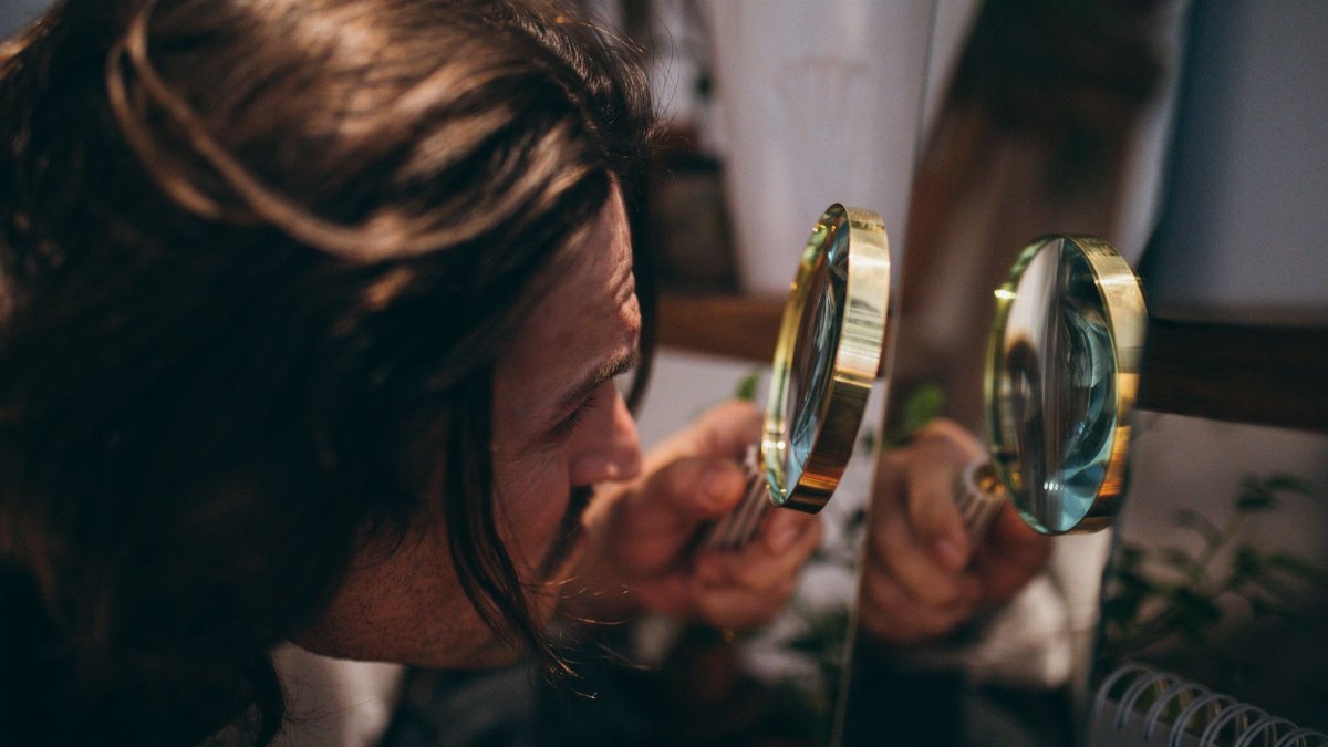 Close-up shot of a man with long hair using a magnifying glass to inspect his reflection in a mirror.