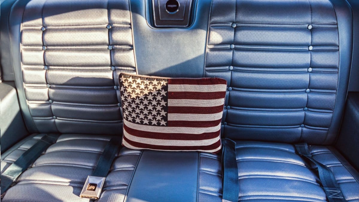 Interior of a vintage blue car with an American flag pillow on the seat, showcasing classic automotive design.