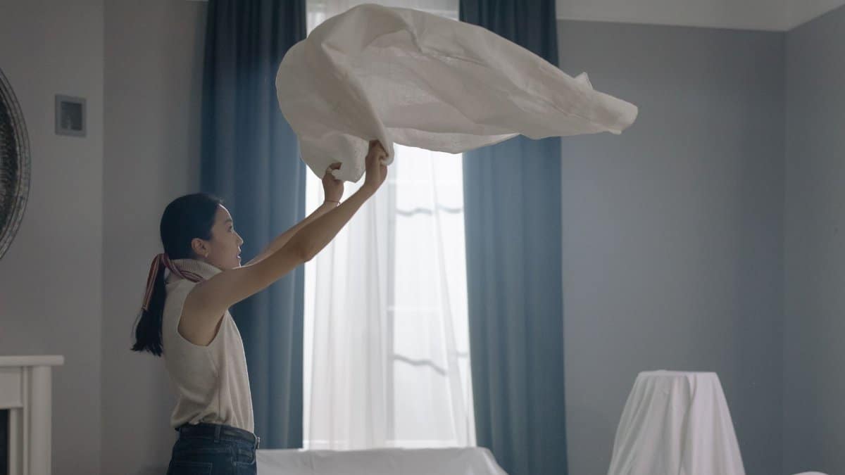 An Asian woman in a sleeveless shirt shakes out a white sheet in a well-lit room.