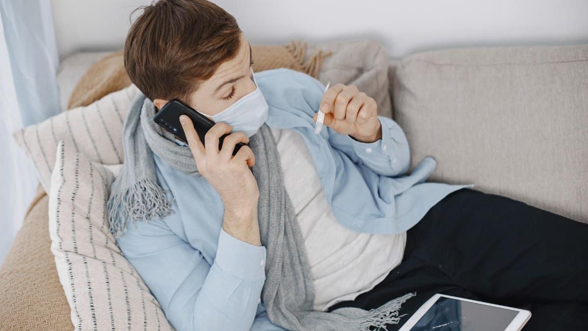 Man using telehealth services while sick at home. Lying on sofa with phone and thermometer.