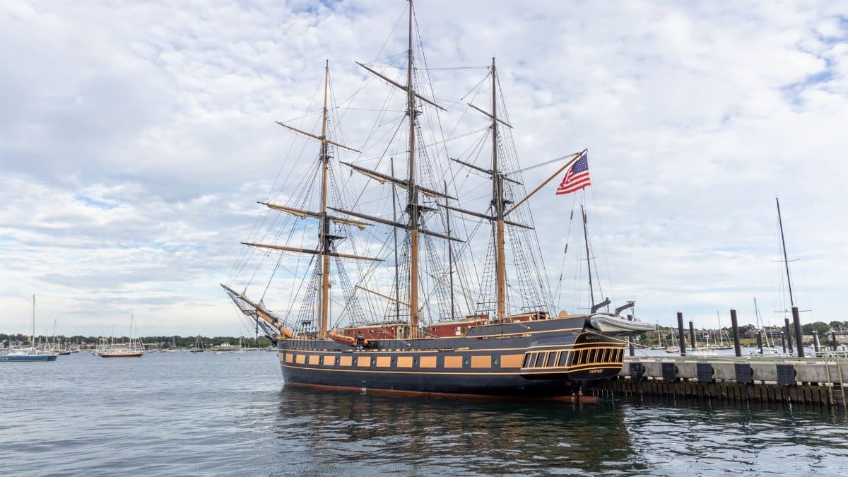 A majestic historic ship with an American flag docked in Newport, RI harbor, under a cloudy sky.