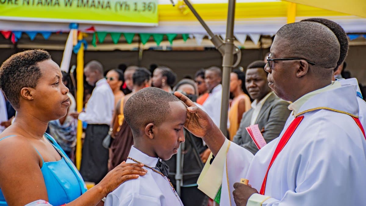 A boy receives a blessing during an outdoor religious ceremony with a priest, surrounded by family and guests.