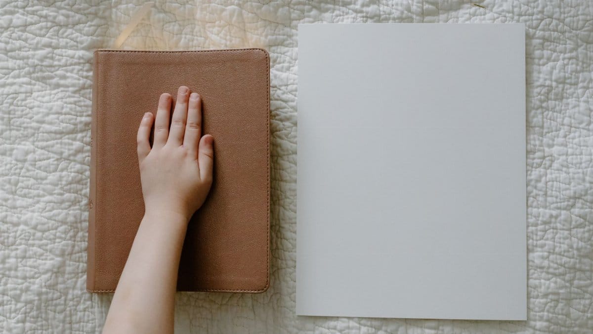 Flat lay of a child's hand on a leather book beside a blank sheet, photographed on a quilt.