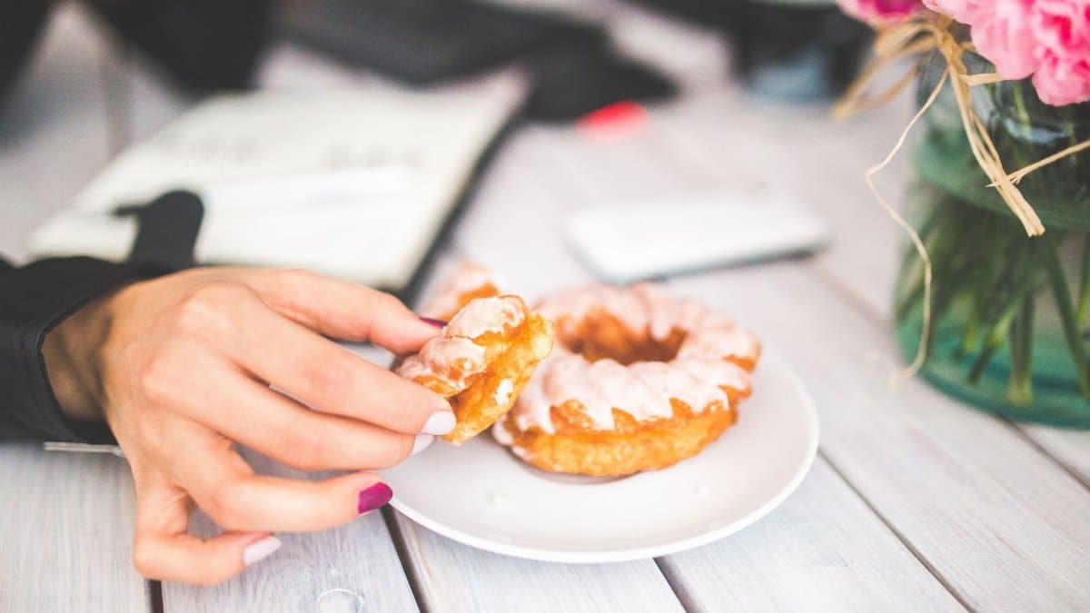 A woman's hand reaches for a glazed donut on a white plate indoors, showcasing a sweet treat moment.