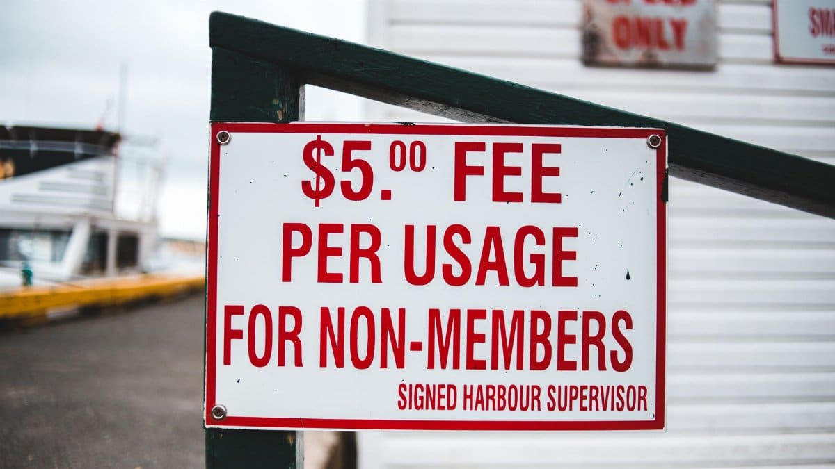 White rectangular sign with red letters informing about harbor entrance fee placed on wooden fence beside waterfront building