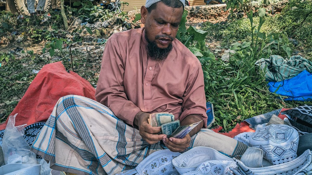 A local vendor in Habiganj, Bangladesh counts money surrounded by traditional hats at an outdoor market.
