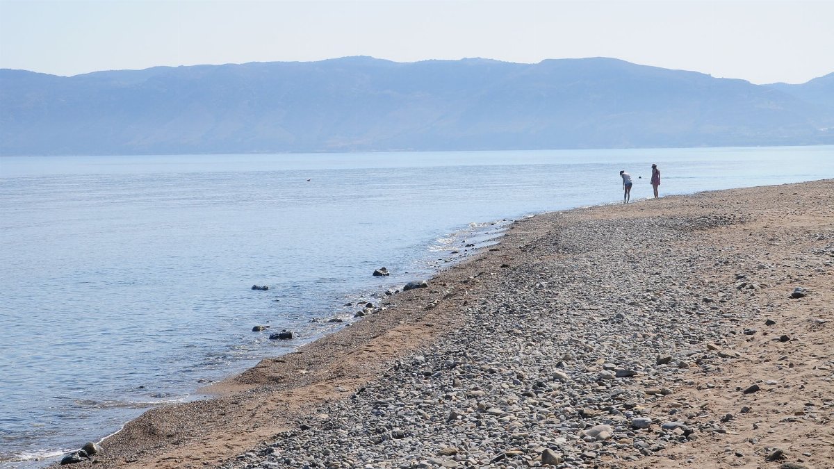 Peaceful beach scene with two people on a rocky shore against a mountain backdrop.