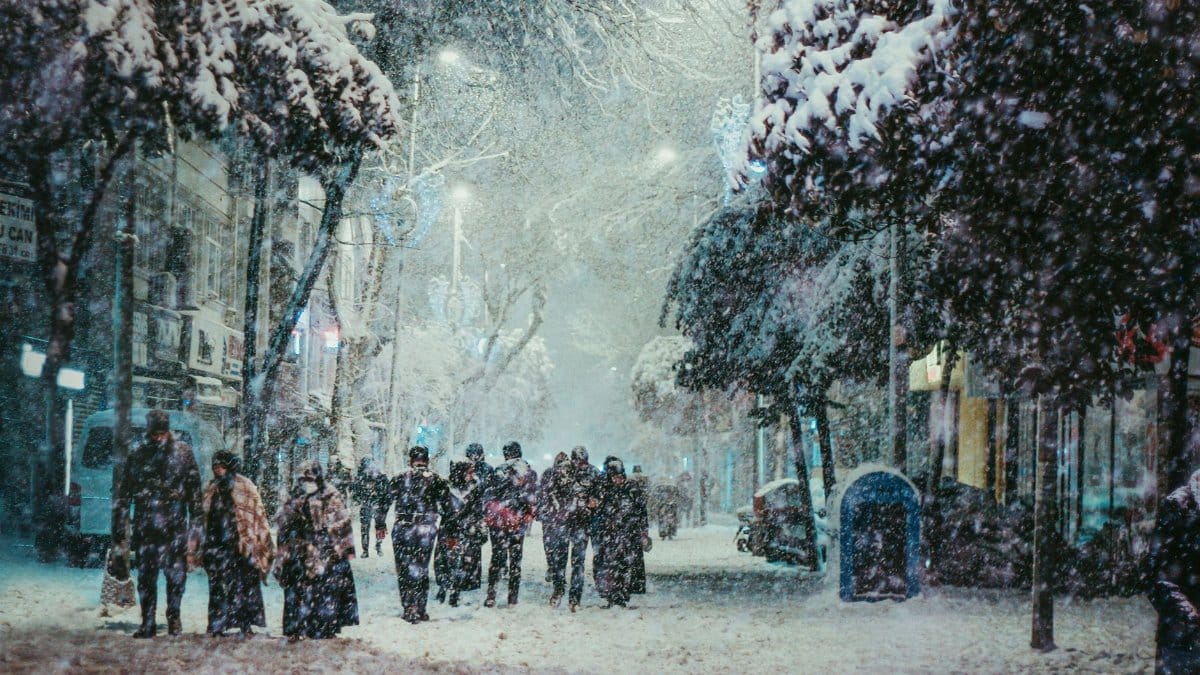 A group of people walking through a snow-covered city street, illuminated under streetlights during winter.