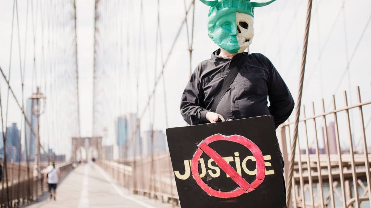 Individual wearing a Statue of Liberty mask holding a justice protest sign on Brooklyn Bridge.