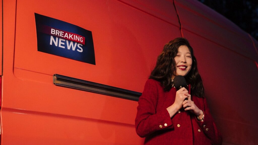 Asian female reporter smiling with microphone besides a news van at night.