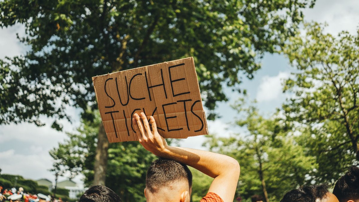 A man in a crowd holds a sign reading 'Suche Tickets' at an outdoor event, seeking extra admissions.