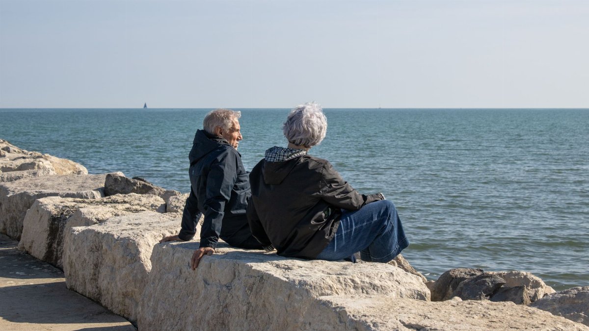 An elderly couple sitting on rocky shore, enjoying the calm sea under the clear sky.