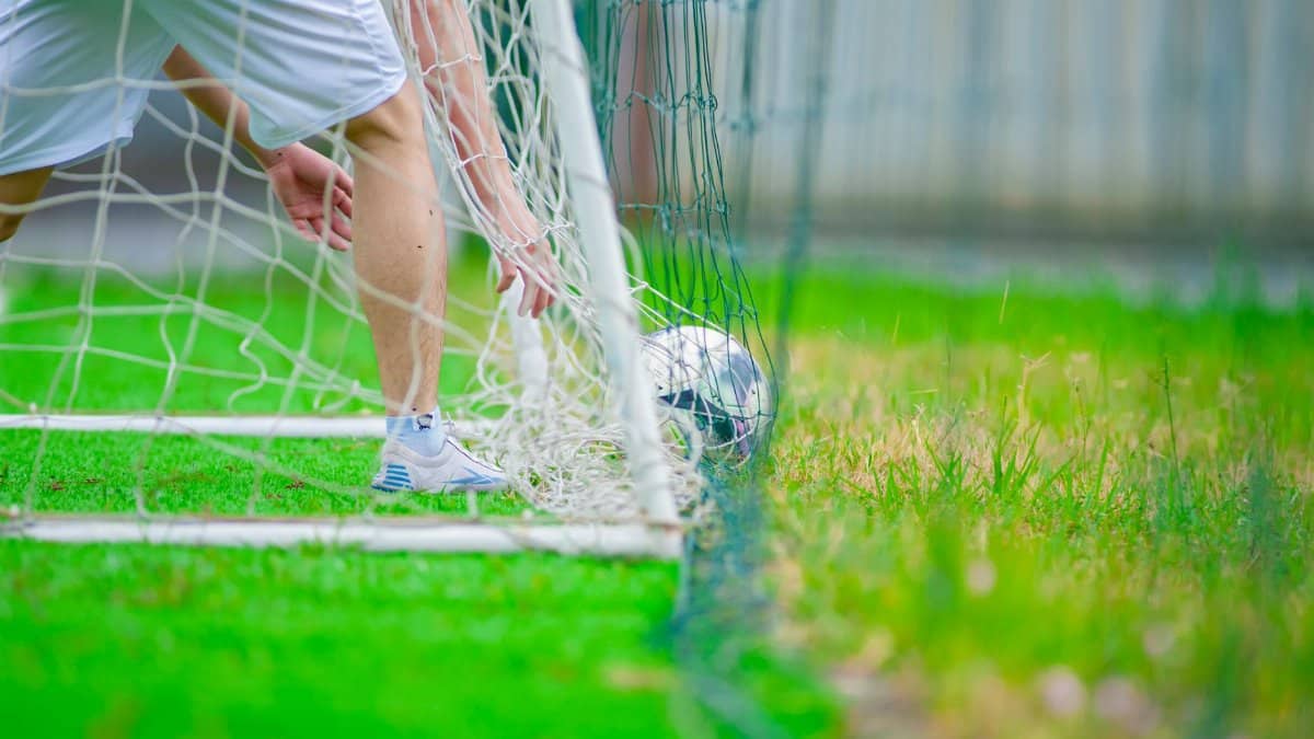 A soccer player retrieves a ball from the goal on a lush green field, captured in Hanoi, Vietnam.