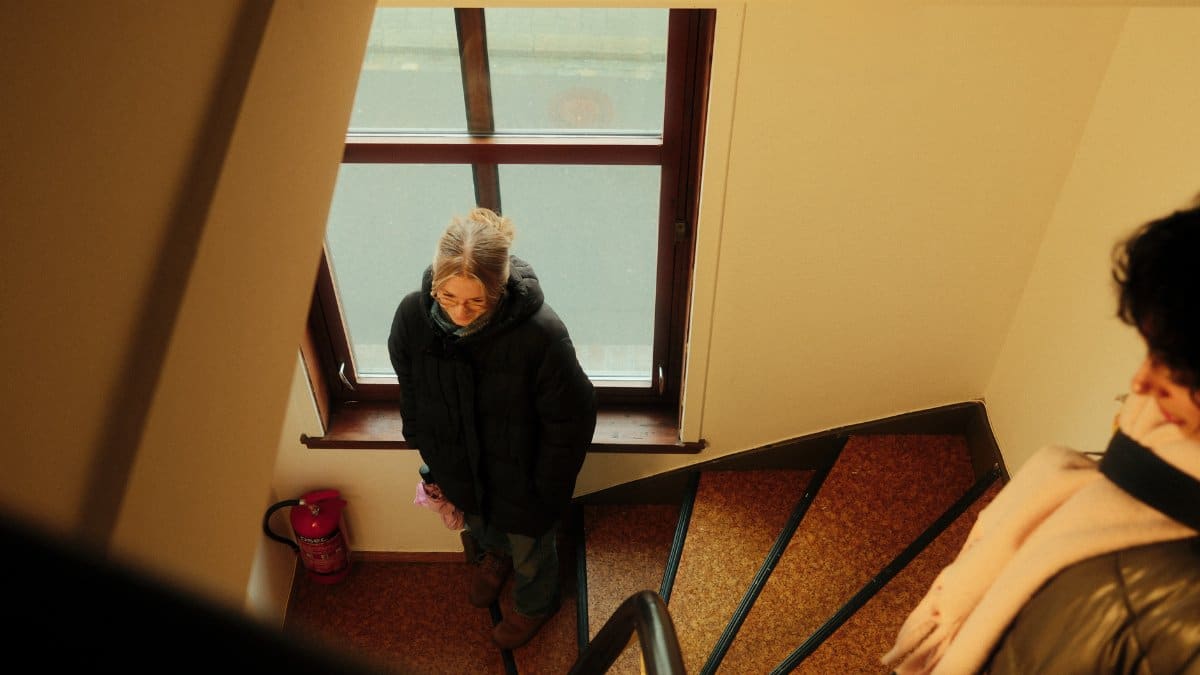 Two adults in winter clothing standing in a warmly lit stairway beside a window.