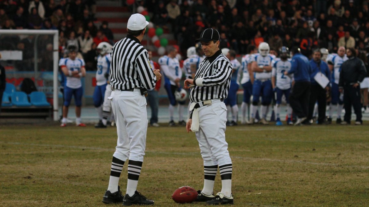 Two referees on a football field preparing for a game with players in the background at a stadium.