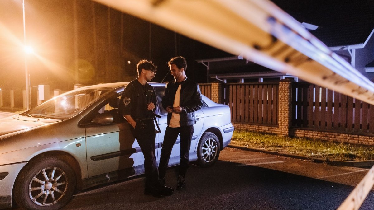 Two men discussing at a nighttime urban crime scene with police tape and a car.