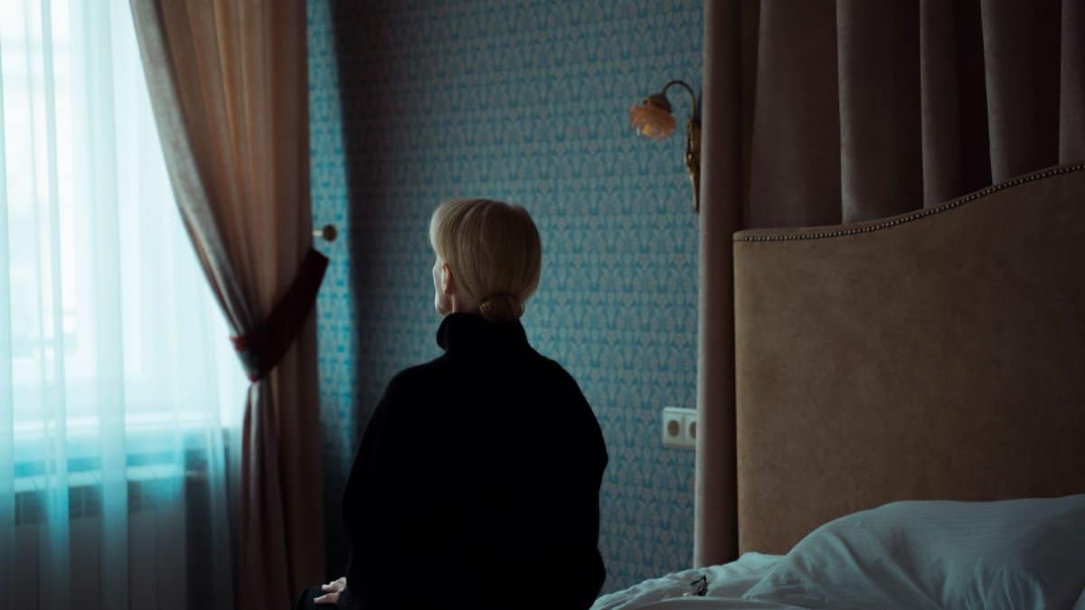 A woman in a dark room sits on a bed facing the window, reflecting solitude and contemplation.