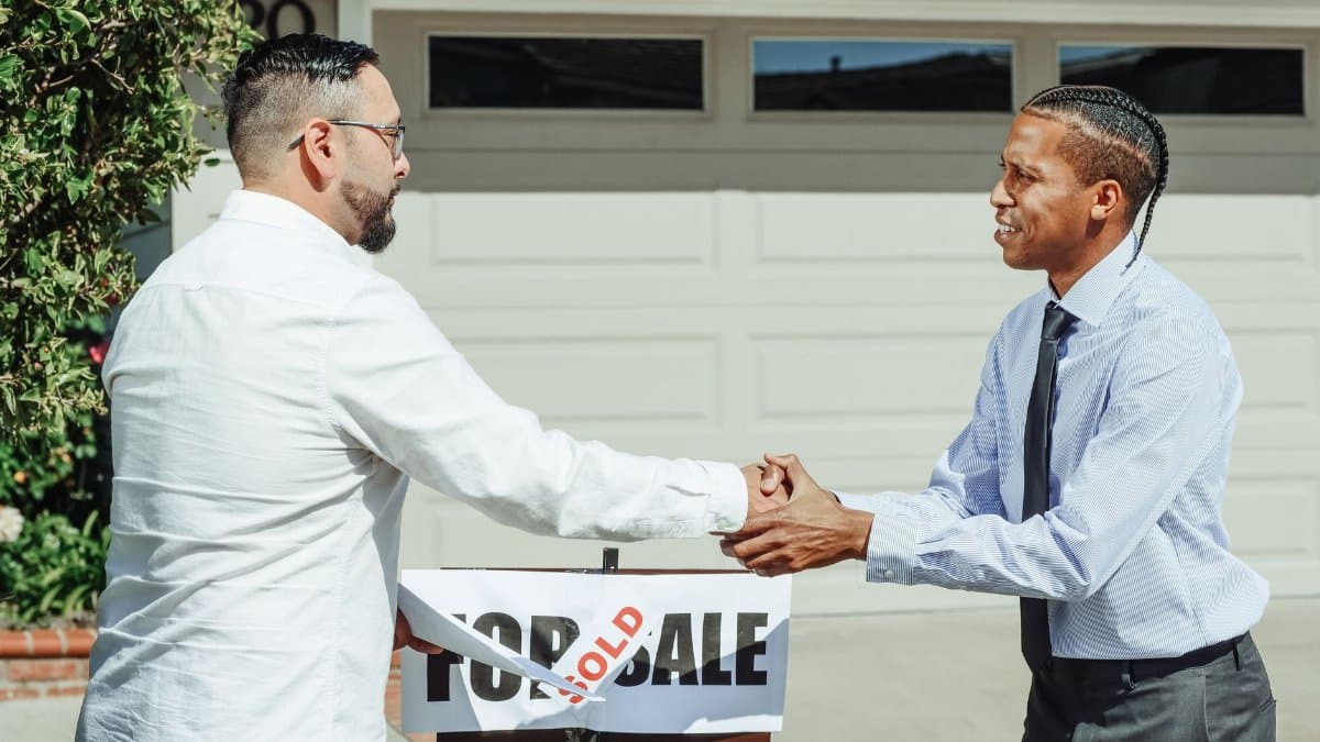 Two men shaking hands in front of a sold house sign, sealing a real estate transaction.
