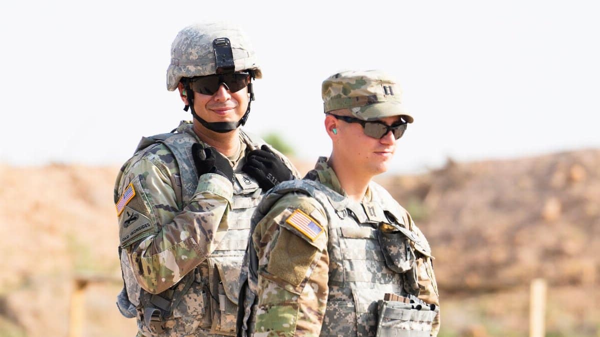 Two US military personnel in camouflage uniforms wearing helmets and sunglasses in a desert setting.