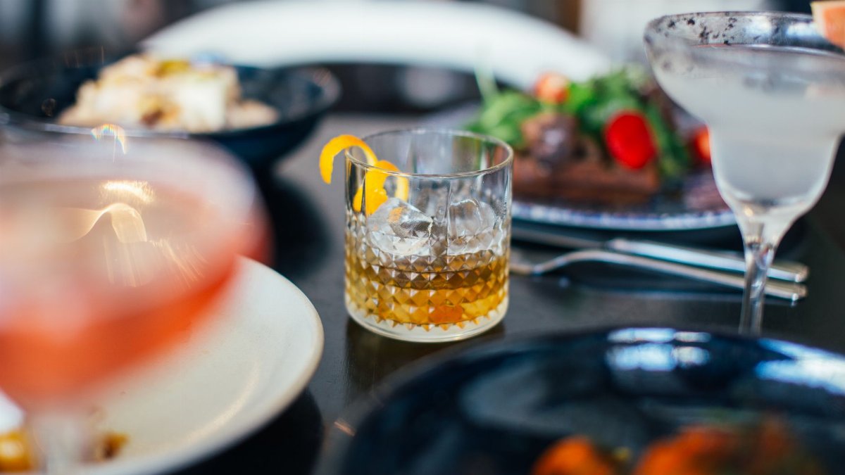 From above glasses of whiskey with ice cubes and lemon peel and vermouth with grapefruit placed on table near assorted dishes in restaurant