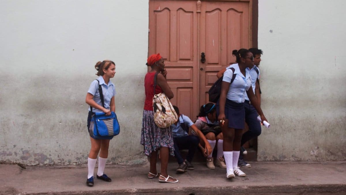 A group of students in their school uniforms standing and sitting on a city sidewalk by a wooden door.