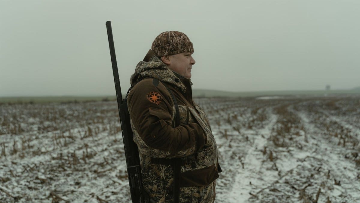 A man stands in a snowy field holding a shotgun, dressed in camouflage gear.