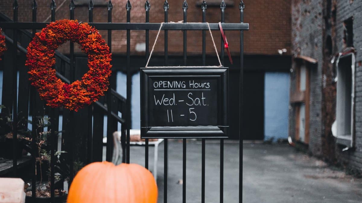 Fall-themed outdoor sign with pumpkins and wreath on metal fence showing business hours.