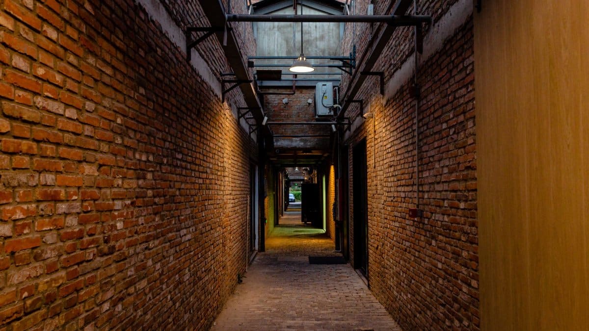 Narrow alleyway with brick walls illuminated by warm lights, leading to an open area.