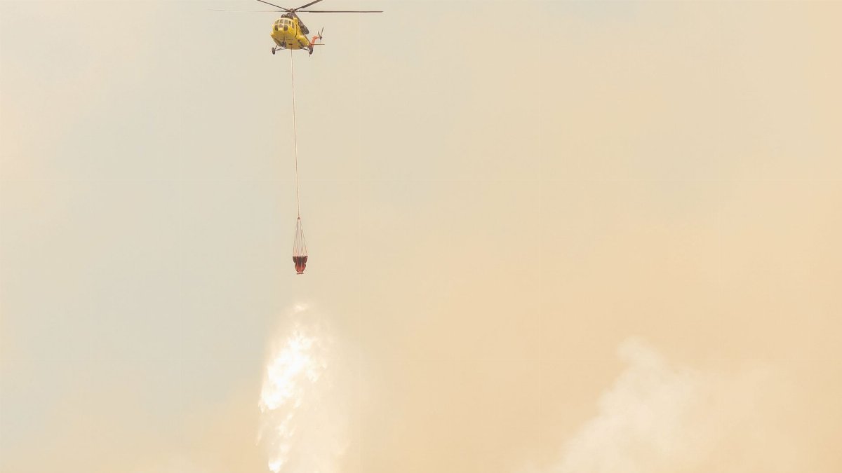 Yellow helicopter drops water to combat wildfires amidst smoky skies.