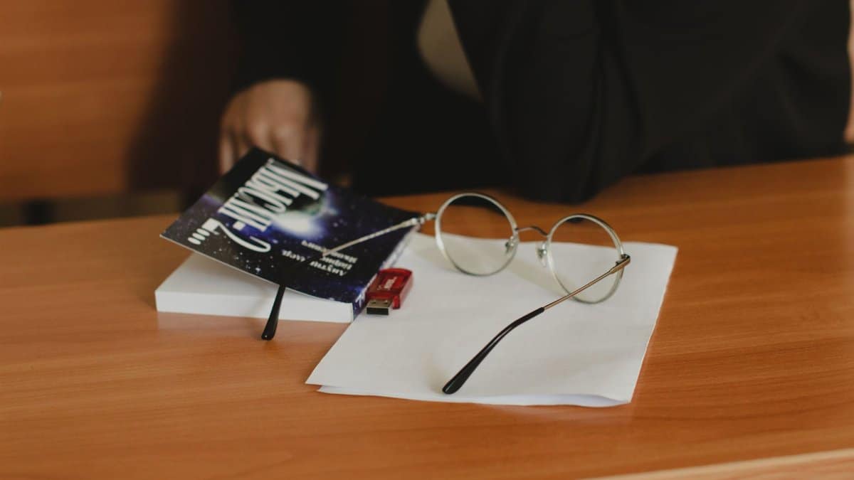 Close-up of eyeglasses, book, and USB drive on a wooden desk indoors.