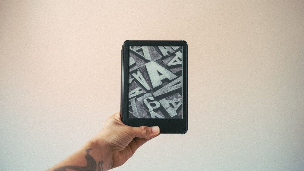 A tattooed hand holding an e-reader displaying typography design against a plain background.