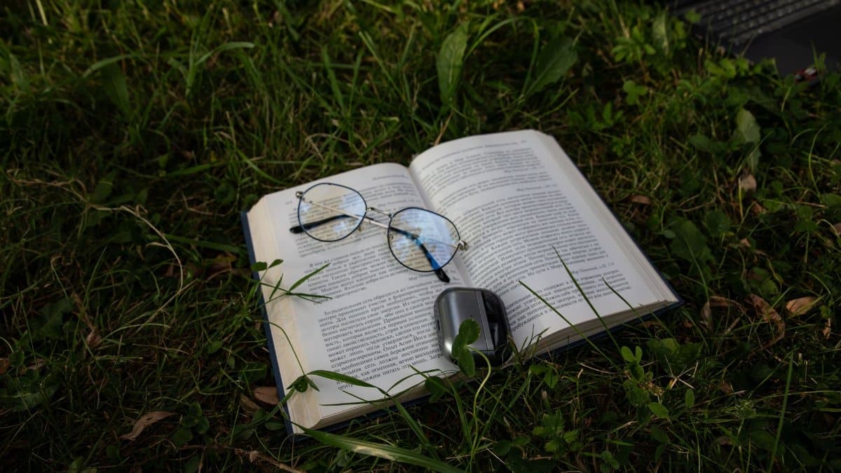 An open book with reading glasses and a mouse on grass, next to a laptop on a sunny day.