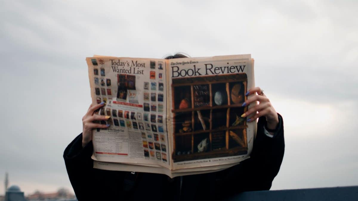 Individual holding a book review newspaper with colorful nails against a cloudy sky backdrop.